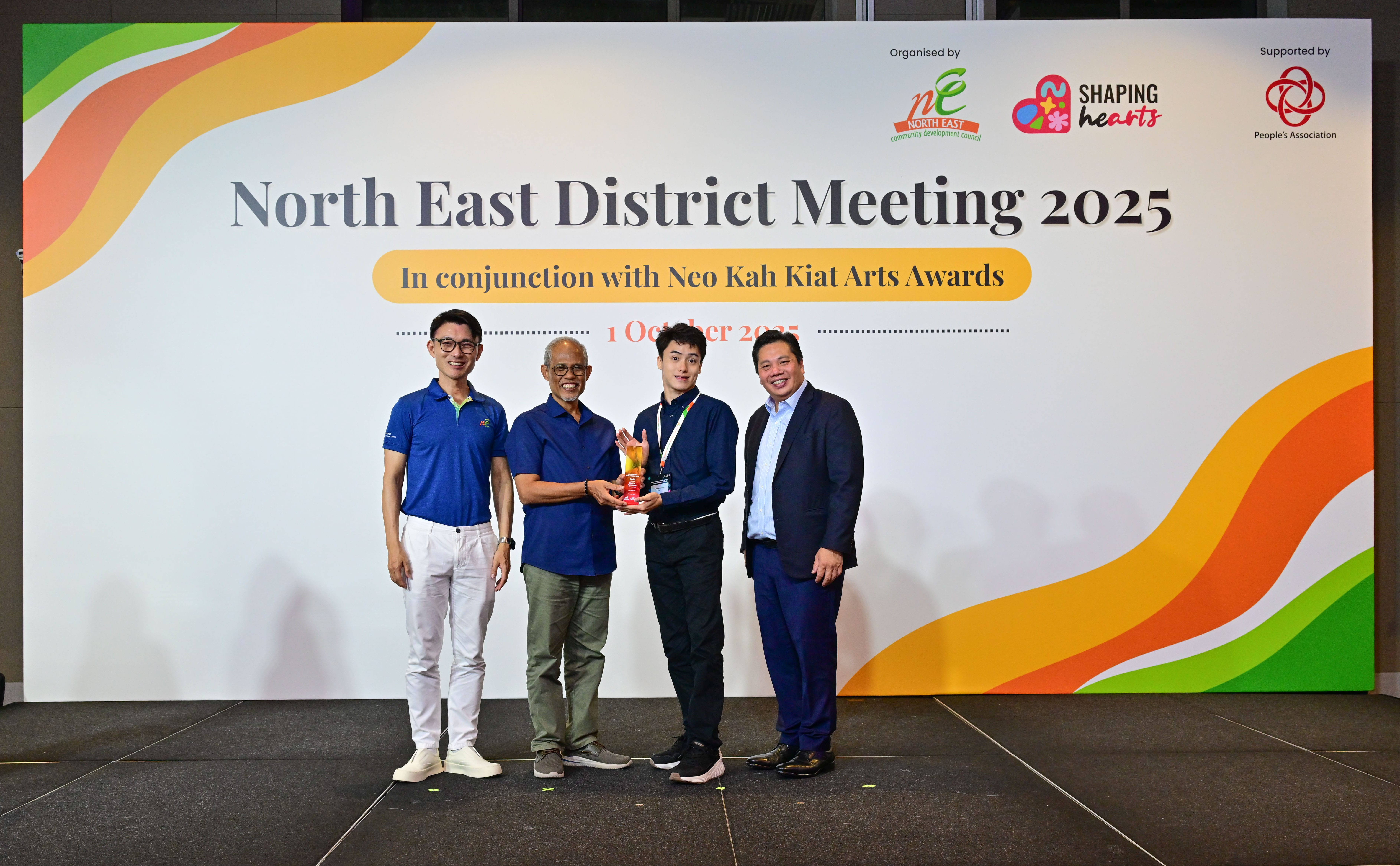 Award recipient standing on stage with the presenter, holding a acrylic plaque and posing for a group photo during the award ceremony, with a backdrop reading ‘North East District Meeting 2025 In conjunction with Neo Kah Kiat Arts Awards'